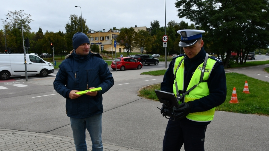 policjant z pracownikiem WORD w Białymstoku trzymającym opaski odblaskowe koloru seledynowego, obsługuje kontroler kamery drona